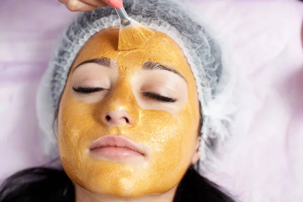 Closeup ένα θηλυκό πελάτη ένα σαλόνι ομορφιάς με μια χρυσή μάσκα στο πρόσωπο. Κοσμετολογία και δέρμα φροντίδα ρουτίνας. — Φωτογραφία Αρχείου