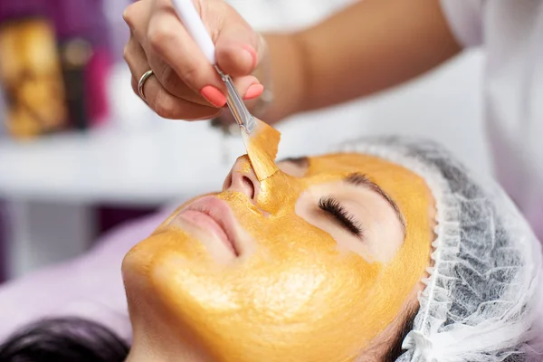 Макрофотография руки косметолога с помощью специальной кисти накладывает на лицо девушки золотую маску в спа-салоне — стоковое фото