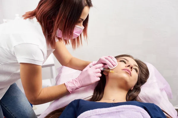 Donna che viene iniettata nel salone di cosmetologia. Medico in guanti medici e maschera con siringa inietta guance farmaco. Iniezioni di bellezza e cosmetologia — Foto Stock