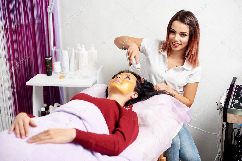 Smiling cosmetologist makes the procedure Microcurrent therapy on the hair of a beautiful, young woman in a beauty salon.