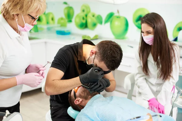 Junger männlicher Zahnarzt behandelt einen männlichen Kunden einer modernen Zahnarztpraxis, während weibliche Berufstätige und junge brünette Assistentinnen dies beobachten. — Stockfoto