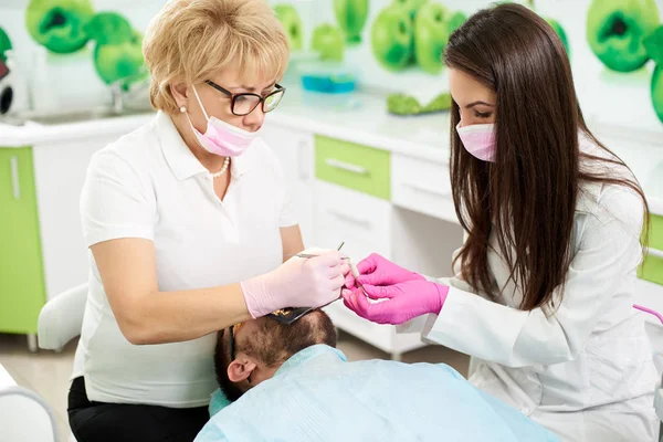 Zahnärztin und ihre junge brünette Assistentin behandeln einen männlichen Kunden einer modernen Zahnmedizin. — Stockfoto