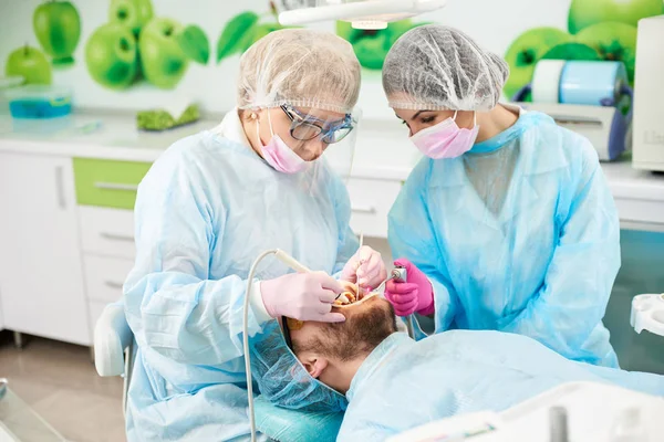 Dois dentistas com vestes médicas azuis estão a encher os dentes de um cliente masculino de um consultório odontológico moderno. . — Fotografia de Stock