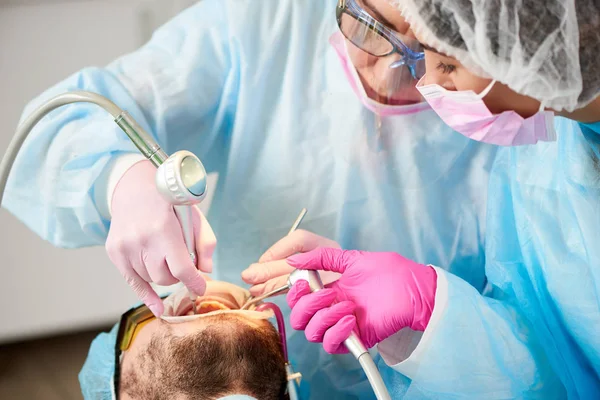 Nahaufnahme einer Zahnärztin und ihrer Assistentin bei der einmaligen Arbeit mit einem männlichen Patienten in einer Klinik mit innovativen Technologien. — Stockfoto