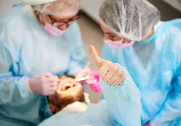 Фотография сверху женщины-врача и ее ассистентки, одетые в одноразовую униформу, работают с пациентом-мужчиной, который показывает класс жестов в современном стоматологическом кабинете — стоковое фото