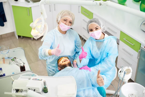 Glückliche und lächelnde Zahnärzte und ihr männlicher Klient zeigen nach erfolgreicher Zahnbehandlung in einer Zahnarztpraxis alle Daumen nach oben. — Stockfoto