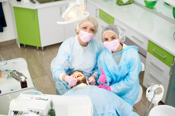 Фото зверху жінки-лікаря та її помічника, одягнені в одноразову однорідну роботу з пацієнтом чоловічої статі в сучасному стоматологічному кабінеті та показують клас жестів — стокове фото