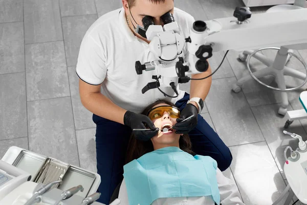 Erkek asistan siyah bir maske ve eldiven dişçiyle bir mikroskop kullanarak bir hasta muamelesi yapıyor. Yeni teknolojilerin kullanımı ile modern diş hekimliği