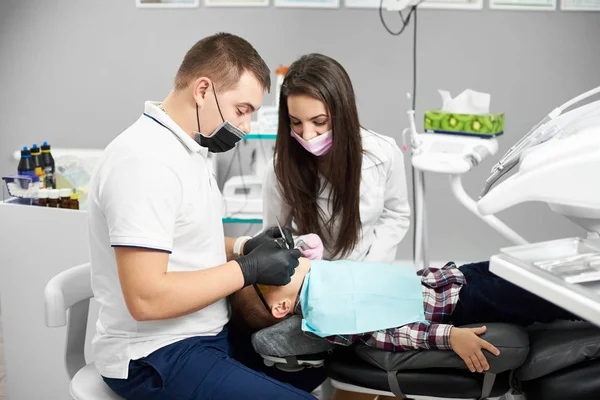 Young male dentist in a white T-shirt with a woman assistant helping him is giving dental treatment to a child in a modern dental clinic