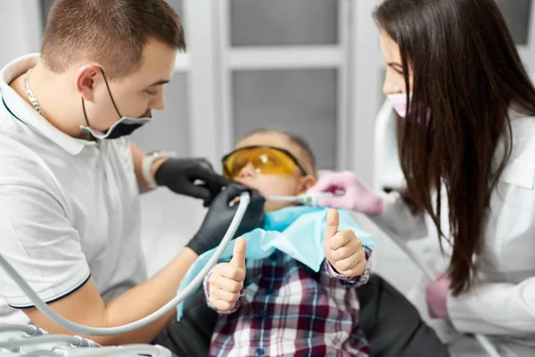 Ein Kind mit orangefarbener Zahnbrille reckt die Daumen in die Höhe, während ein männlicher Zahnarzt in blauem T-Shirt und schwarzen Handschuhen zahnärztliche Behandlung durchführt, wobei ihm eine brünette Assistentin hilft. — Stockfoto