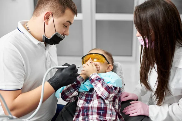 Ein kleiner Junge im Zahnarztstuhl schließt seinen Mund mit den Händen, aus Angst vor einem Zahnarzt, der eine Bohrmaschine in der Hand hält. moderne Zahnklinik — Stockfoto