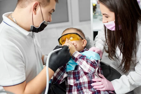Ein Kind mit orangefarbener Zahnbrille verdeckt erschrocken seinen Mund, bevor seine Zahnbehandlung von einem jungen Zahnarzt und seiner attraktiven Assistentin durchgeführt wird. — Stockfoto