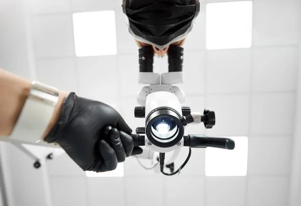 Upwardview manliga tandläkare med svart gummihandskar titta i mikroskopet med hjälp av professionella Dentalutrustning i en vit ljusa tandläkare kontor. — Stockfoto