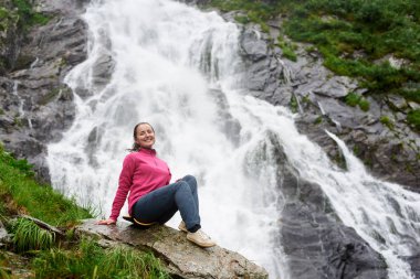 Mutlu kadın Fagarash dağlar boşaltmak güç doğa sağlık mutluluk yaşam tarzı aktivite spor hiking, kamp içinde güzel bir Balea şelale önünde oturan kameraya gülümseyen.