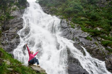 Genç kadın bir kayaya elleriyle şelale Balea Fagarash dağlarda önünde oturup. Karpatlar Romanya, Transfagarasan road yakınındaki yer.