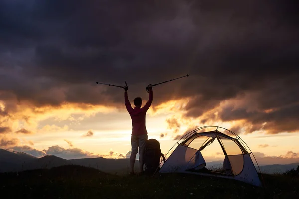 Reaching the top. Toned young female enjoying beautiful sunset with walking sticks in her hands near tent on top of mountain hill. Night evening tourist climber majestic amazing view scenery — Stock Photo, Image