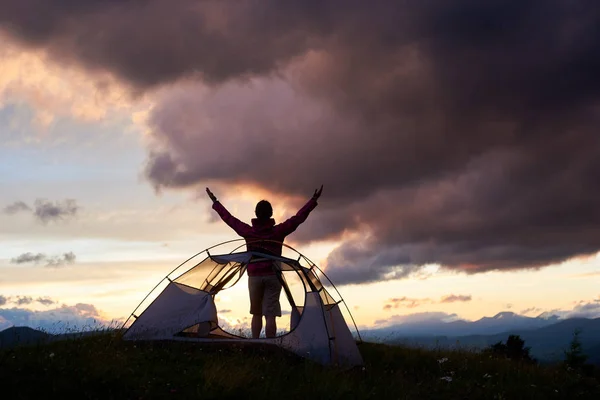 Happy tourist woman standing near the tent with raised up hands enjoying view of mountains. Travel to wild nature, happiness emotion, summer holidays, unity with nature concept. — Stock Photo, Image