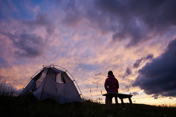 Mujer excursionista sentada en un banco cerca de una tienda de campaña y mirando a la increíble vista del cielo de la noche puesta de sol con nubes púrpura. Joven turista sola en clima frío cerca del camping al aire libre. Concepto de viaje . — Foto de Stock