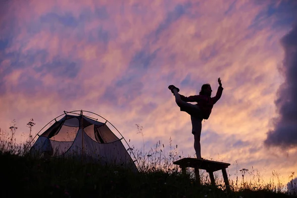 Joven hermosa mujer practica yoga en una caminata cerca de la tienda. Estilo de vida activo, viajes, vacaciones, vacaciones y concepto de aventura. Silueta de una chica de pie sobre una pierna en un banco al atardecer . — Foto de Stock