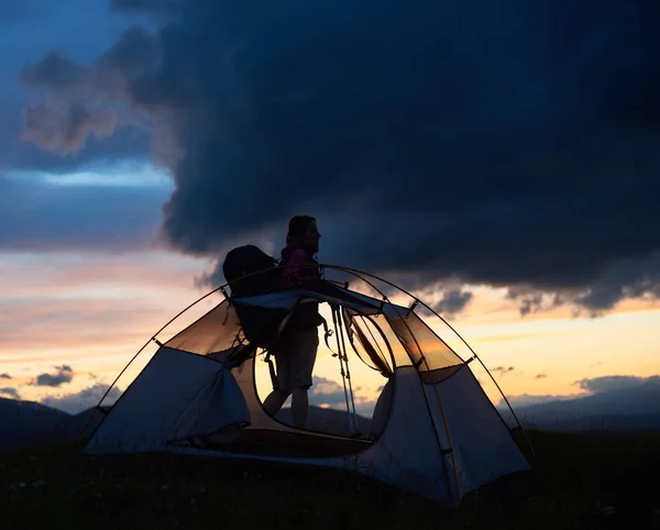 Silueta de una joven senderista cansada con bastones de trekking y una mochila cerca de la tienda, hora de la puesta del sol con el sol naciente en las montañas — Foto de Stock