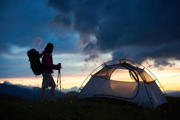 Silhueta de turista feminina perto da tenda com mochila nos ombros e bengalas nas mãos desfrutando da vista do pôr do sol sobre as colinas da montanha. Viagens e conceito ativo de modo de vida . — Fotografia de Stock
