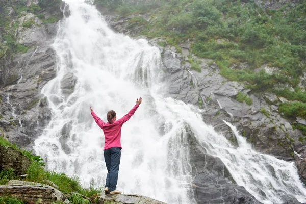 Imagen de la espalda de una turista levantando las manos disfrutando de una hermosa vista impresionante de la cascada de Balea en Rumania. Mujer chica agua verde roca increíble maravillosa naturaleza espectacular paisaje — Foto de Stock