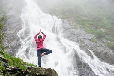 Genç ve güzel kız büyük güçlü şelale yakınındaki bir yoga poz ayakta. Güzel kadın sakin vahşi doğada meditasyon. Şelale Balea Romen Fagarash dağlarında seyahat.