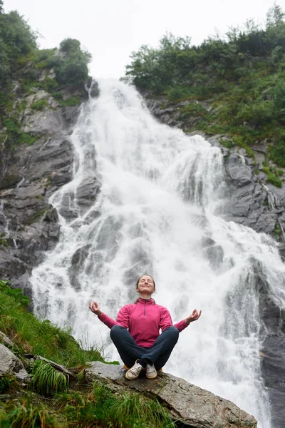 Щаслива жінка, сидячи в позі лотоса біля водоспаду, роздумуючи з закритими очима копія — стокове фото