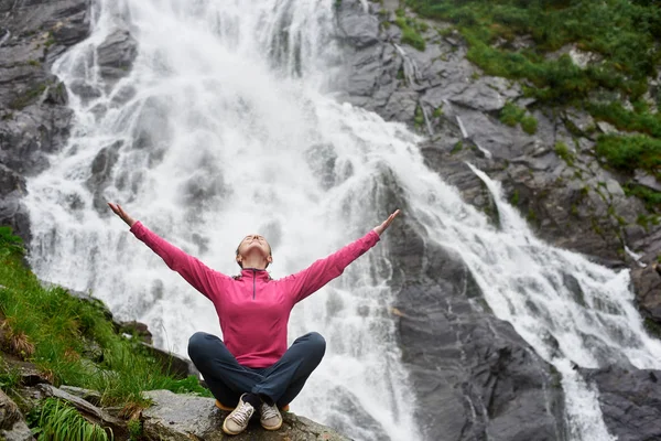 Turista mujer caucásica se sienta con los brazos abiertos y mira hacia arriba con deleite contra el fondo de la cascada Balea en Rumania. Cascada rocosa en las montañas . — Foto de Stock