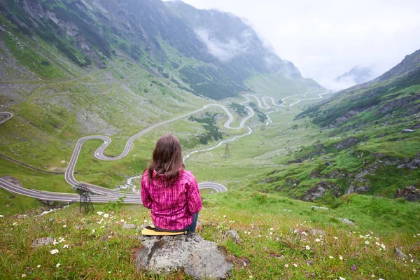 Tânăra brunetă, așezată pe piatră și admirând priveliștea spectaculoasă a rocilor verzi și a pajiștilor și a drumului transagarashan din România. Femeie alpinist turist fericit liniște munți — Fotografie, imagine de stoc