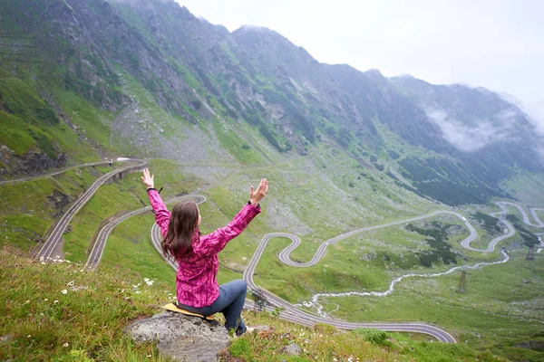 Tânăra femeie cu mâinile sus admirând frumusețea rocilor verzi și a munților și una dintre cele mai populare atracții turistice din lume care înfășoară drumul Transfăgărășan din România. Femeie alpinist bucurie — Fotografie, imagine de stoc