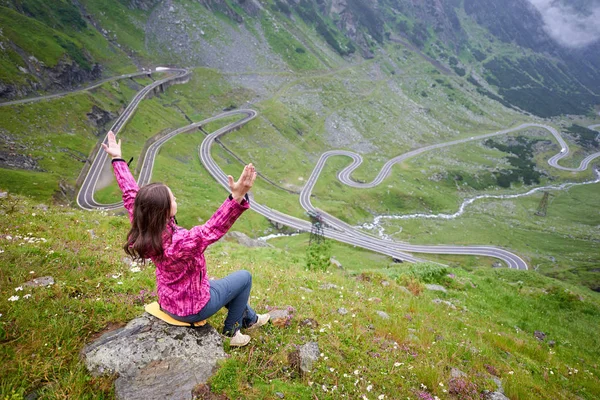 Joven mujer levantando las manos al cielo admirando la belleza de rocas verdes y montañas y una de las atracciones turísticas más populares del mundo sinuosa carretera Transfagarashan en Rumania. Mujer trepadora alegría —  Fotos de Stock
