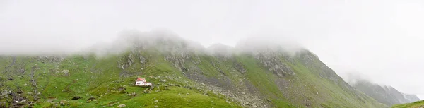 Casă mică și singuratică în munți stâncoși, cu vedere la vale adâncă. Vârfuri muntoase înalte acoperite cu pietre, pietre și nori joși peste coliba turistică din munții Carpați din România . — Fotografie, imagine de stoc