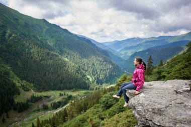 Yeşil çimenli yamaçları ve ağaç, çam ağaçları ve Romanya'da pines dağlar nefes kesen manzaranın rock kenarında oturan gülümseyen güzel kadın turist. Kadın dağcı mutlu şaşırtıcı doğa