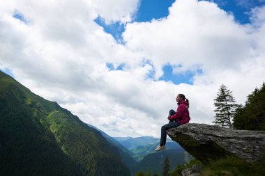 Profile of the girl sitting on the rock, dangling her legs in the abyss against the backdrop of green mountains with forests and clouds above them through which the blue sky is visible. clipart