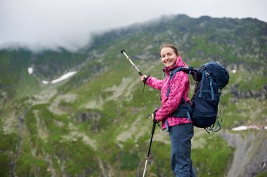 Sisli bulutlu yeşil rocky Dağları cephedeki trekking bir sopayla işaret mavi sırt çantası ile uzun yürüyüşe çıkan kimse kız gülümsüyor. Pembe ceket ve gri pantolon geniş dağ vadinin manzarayı genç kadın.