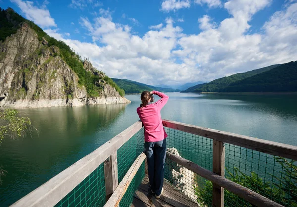 女性摄影师的后方看法在湖 Vidraru 喀尔巴阡山罗马尼亚。阳光明媚的一天美丽的风景: 高山, 森林, 湖泊, 蓝天白云 — 图库照片