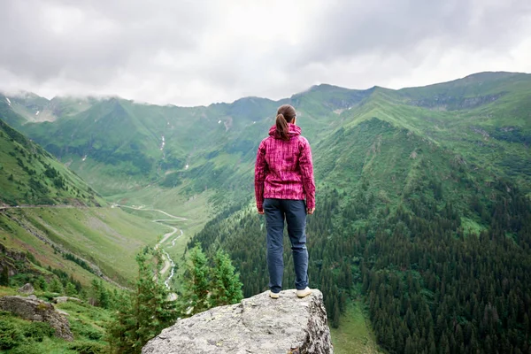 Вид сзади на молодую женщину-путешественницу, стоящую на краю скалы среди великолепных зеленых гор с травянистыми склонами и извилистыми дорогами возле Транскагарашанской дороги в Румынии — стоковое фото