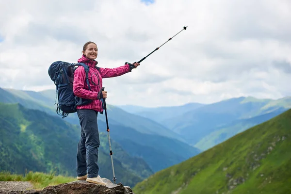 Hiking girl with trekking sticks and a backpack stands on a rock with a blurred background of the mighty mountains of Romania and clouds above them. Girl with trekking stick shows to the distance