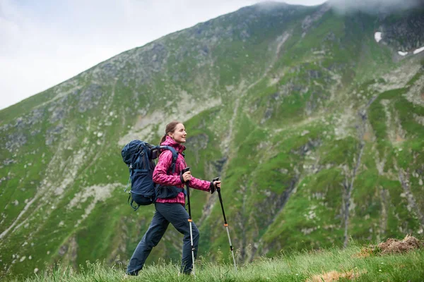 Incroyable touriste féminine marchant sur la pente verte herbeuse avec bâton de marche et sac à dos en face de belles montagnes rocheuses près de la route Transfagarashan en Roumanie. Femme alpiniste vue sur la colline Voyage — Photo