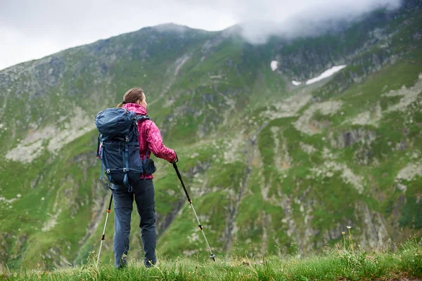 Giovane turista femminile con zaino e bastoni da passeggio ammirando belle montagne rocciose innevate coperte di fitta nebbia in Romania. Donna arrampicatore collina vista spettacolare paesaggio magnifico viaggio — Foto Stock