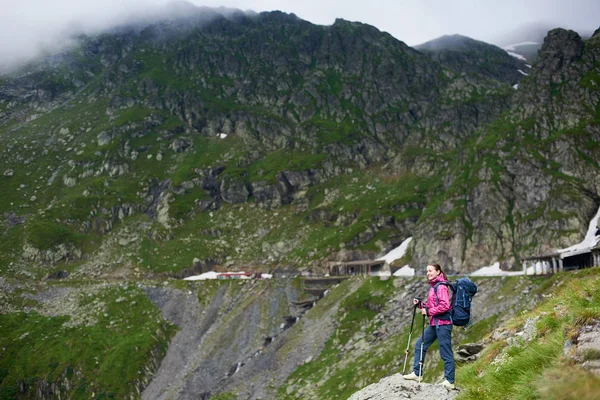 Zâmbind fată drumeție cu rucsac albastru și bastoane de trekking în picioare într-o vale sub munți stâncoși cu nori cețoși. Tineri excursionist femeie în jachetă roz și pantaloni gri care se bucură de vedere la valea muntelui — Fotografie, imagine de stoc