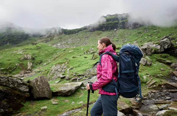 Menina caminhante sorridente com grande mochila azul e varas de trekking andando em verdes montanhas rochosas de outono com nuvens nebulosas. Jovem caminhante mulher em jaqueta rosa desfrutando de vista de grande vale montês . — Fotografia de Stock