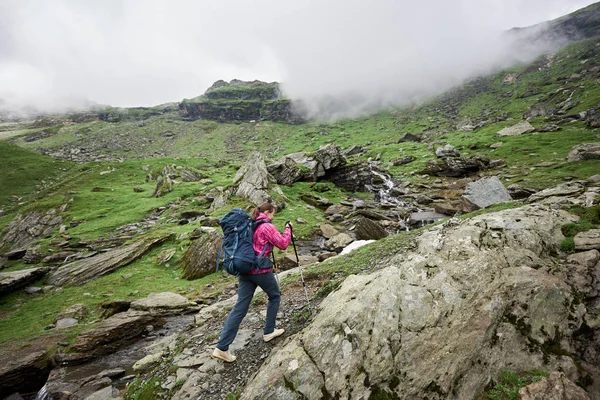 Caminhante menina sobe ao topo do país montanhoso da Romênia. Nuvens embainham montanhas. Mulher com mochila e paus de trekking — Fotografia de Stock