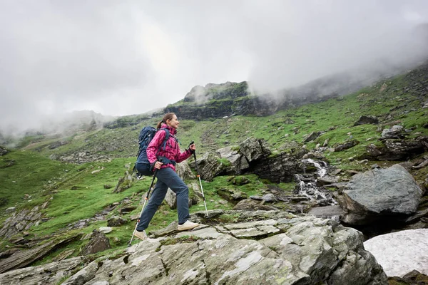 Femme de randonnée souriante avec sac à dos bleu et bâtons de randonnée marchant vers le haut dans les montagnes rocheuses verdoyantes. Jeune randonneuse en veste rose et pantalon gris profitant d'une vue sur la vallée de montagne et les nuages sombres . — Photo