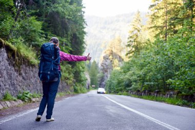 Uzun yürüyüşe çıkan kimse kız yolun kenarında otostop kadar el başparmak ile mavi sırt çantası ile. Genç Gezgin kadın arabayla gezmeye yakalamak istiyor. Backpacker açık yolculuk için hazır.