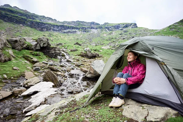 Happy tourist female sits in a tent near a mountain stream in a rocky terrain of Fagaras Mountains. — Stock Photo, Image