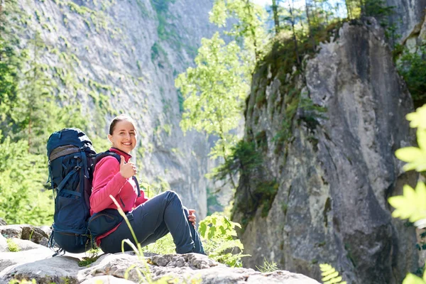 Femeia tânără veselă zâmbind cu bucurie arătând degetele sus stând pe o stâncă odihnindu-se cu rucsacul ei după drumeții în munți copyspace sport activ de viață care călătoresc explorând . — Fotografie, imagine de stoc