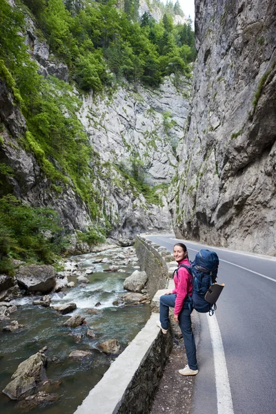 Mujer bonita con mochila en la carretera junto a un arroyo de montaña en la garganta de Bicaz. Cañón es una de las carreteras más espectaculares de Rumania-Cárpatos . —  Fotos de Stock