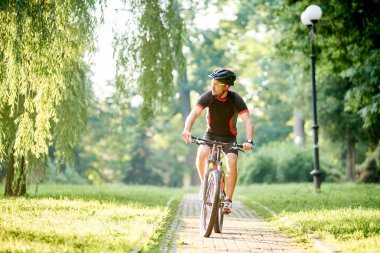 Toned man bicyclist looking to side while riding bicycle down park alley on summer day. Sportsman training thinking about future win in contest. Exercising, reaching goal clipart
